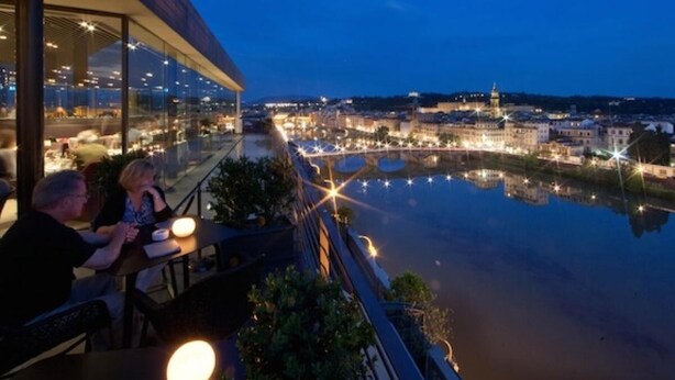 Un área de comedor al aire libre con vistas a un río y al paisaje urbano al anochecer.