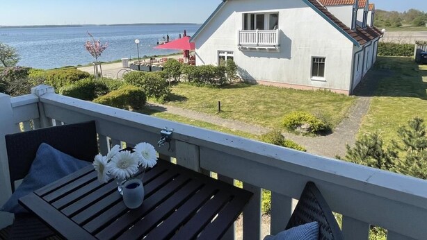 Una casa blanca con techo rojo, un balcón con mesa y flores, y vista al mar.