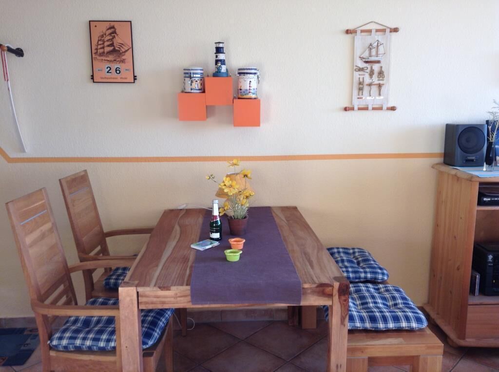 Una mesa de comedor de madera con mantel morado, rodeada de sillas de madera con cojines a cuadros azules y blancos. Hay una pequeña planta en maceta con flores amarillas sobre la mesa.