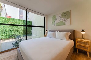 Un dormitorio moderno con un ventanal grande, una cama con ropa blanca, una mesita de noche con lámpara y una planta colgante en la pared.