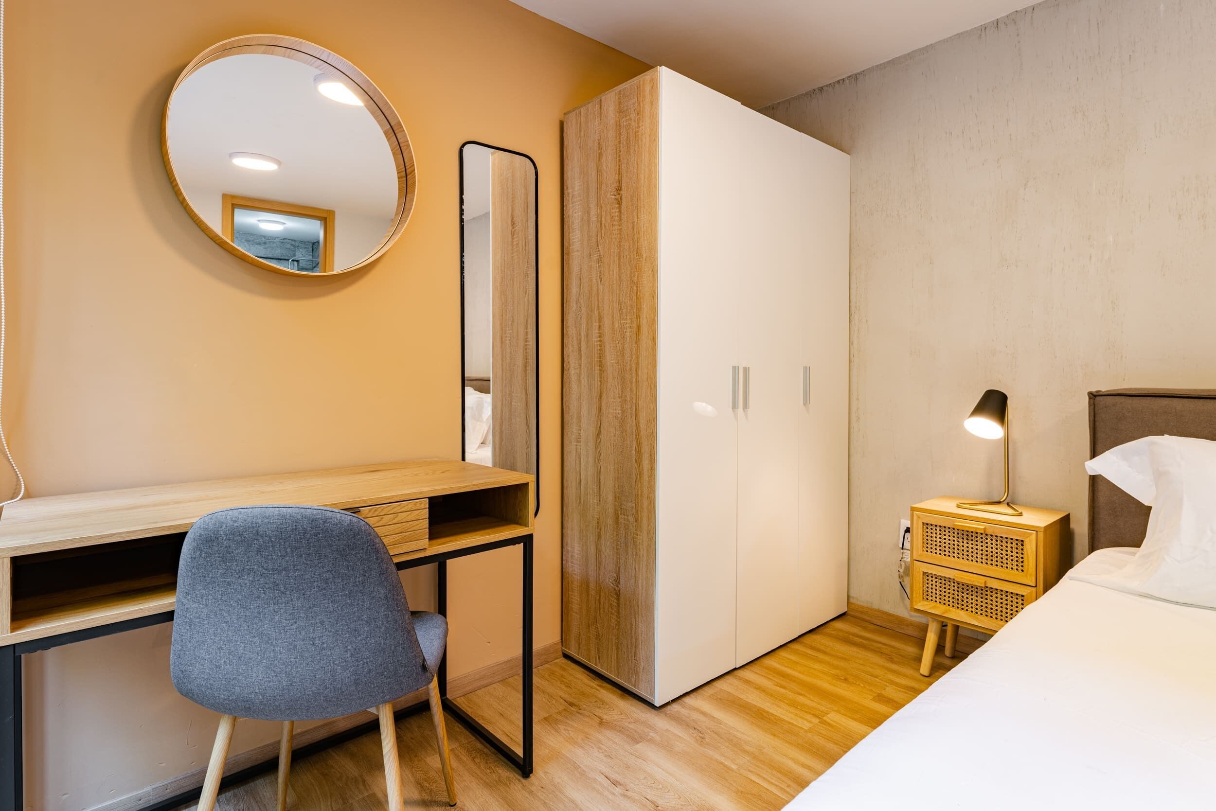 A bedroom with a wooden desk, a chair, a wardrobe, a bedside table with a lamp, and a bed.