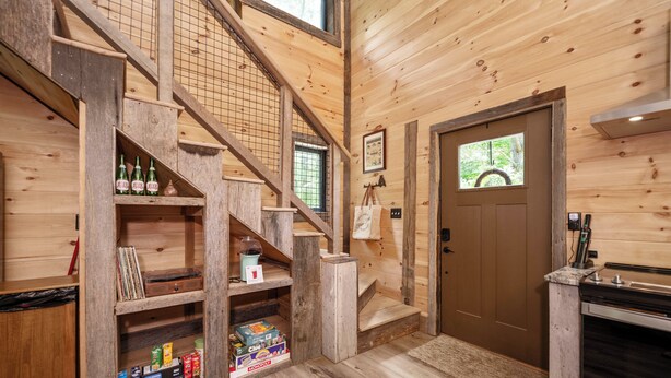 Un intérieur en bois avec un escalier, des étagères remplies de divers objets, une porte et un coin cuisine.