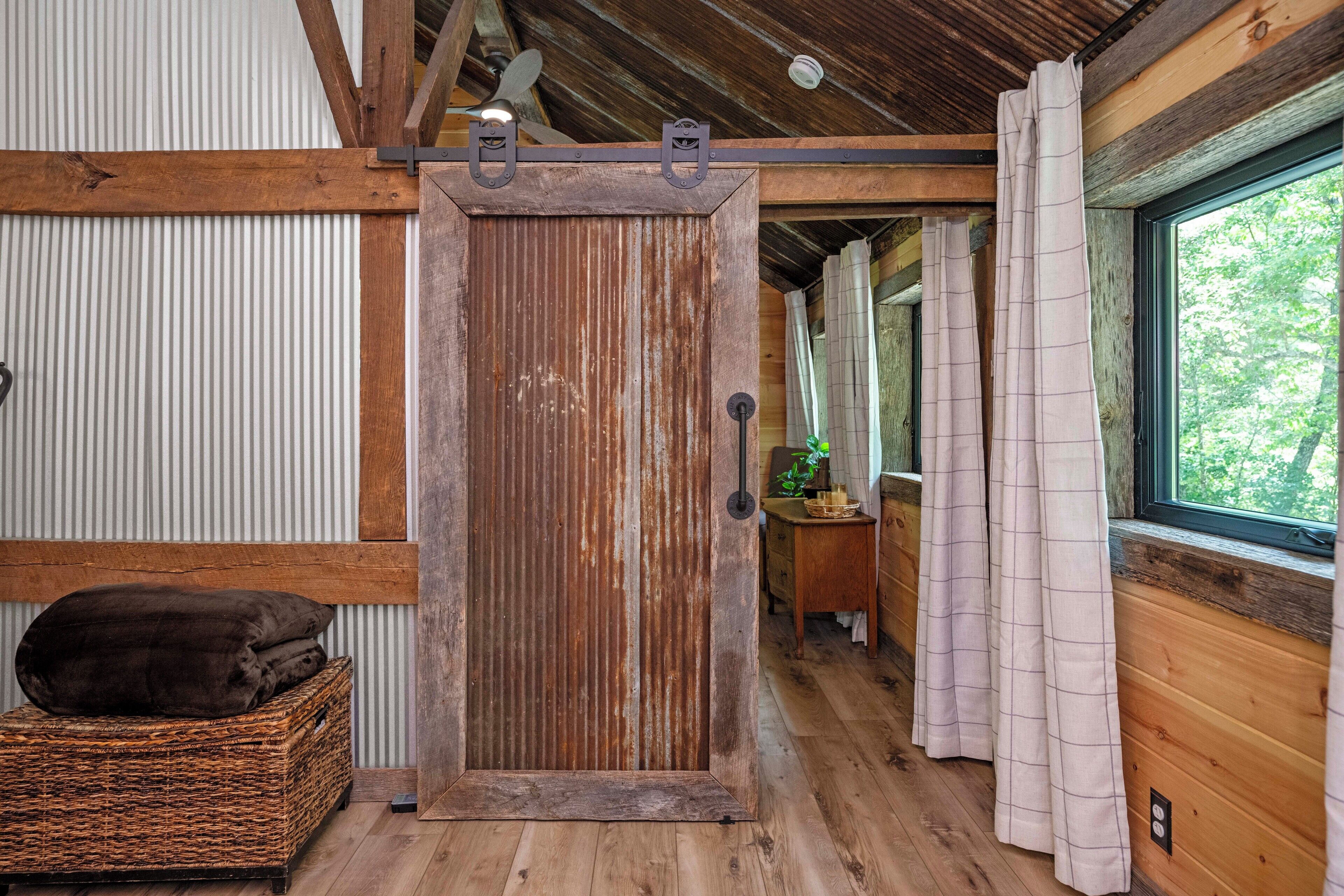 A rustic wooden room with a sliding door, a wicker basket with cushions, a wooden dresser, and a window with curtains.