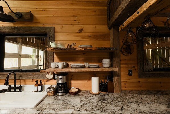 A wooden cabin kitchen with a sink, coffee maker, shelves with dishes, and hanging light fixtures.