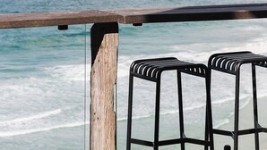 A wooden railing overlooking a beach with waves, and two black metal barstools in the foreground.
