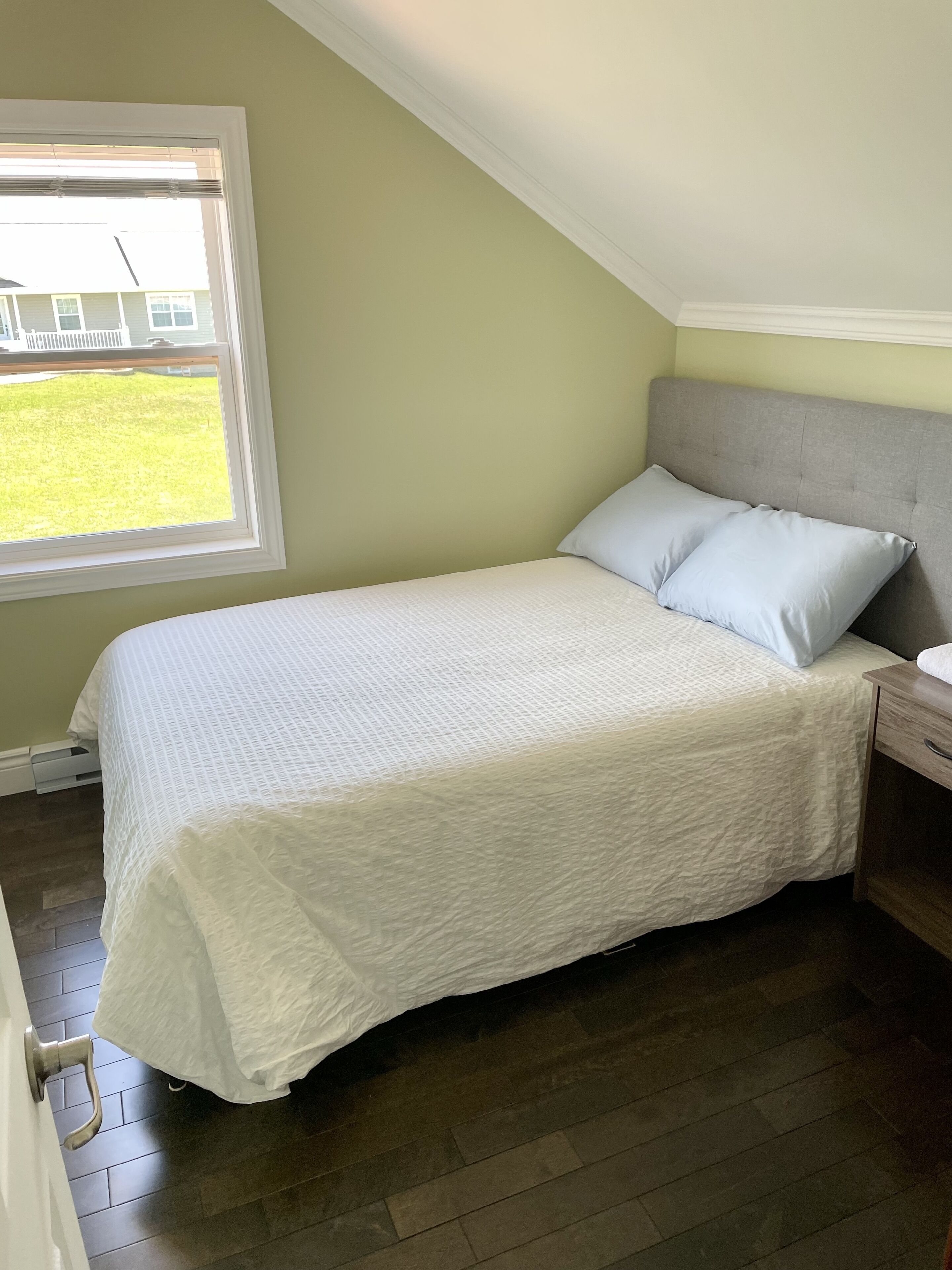 A bedroom with a bed, a nightstand, a window, and a view of the outside.
