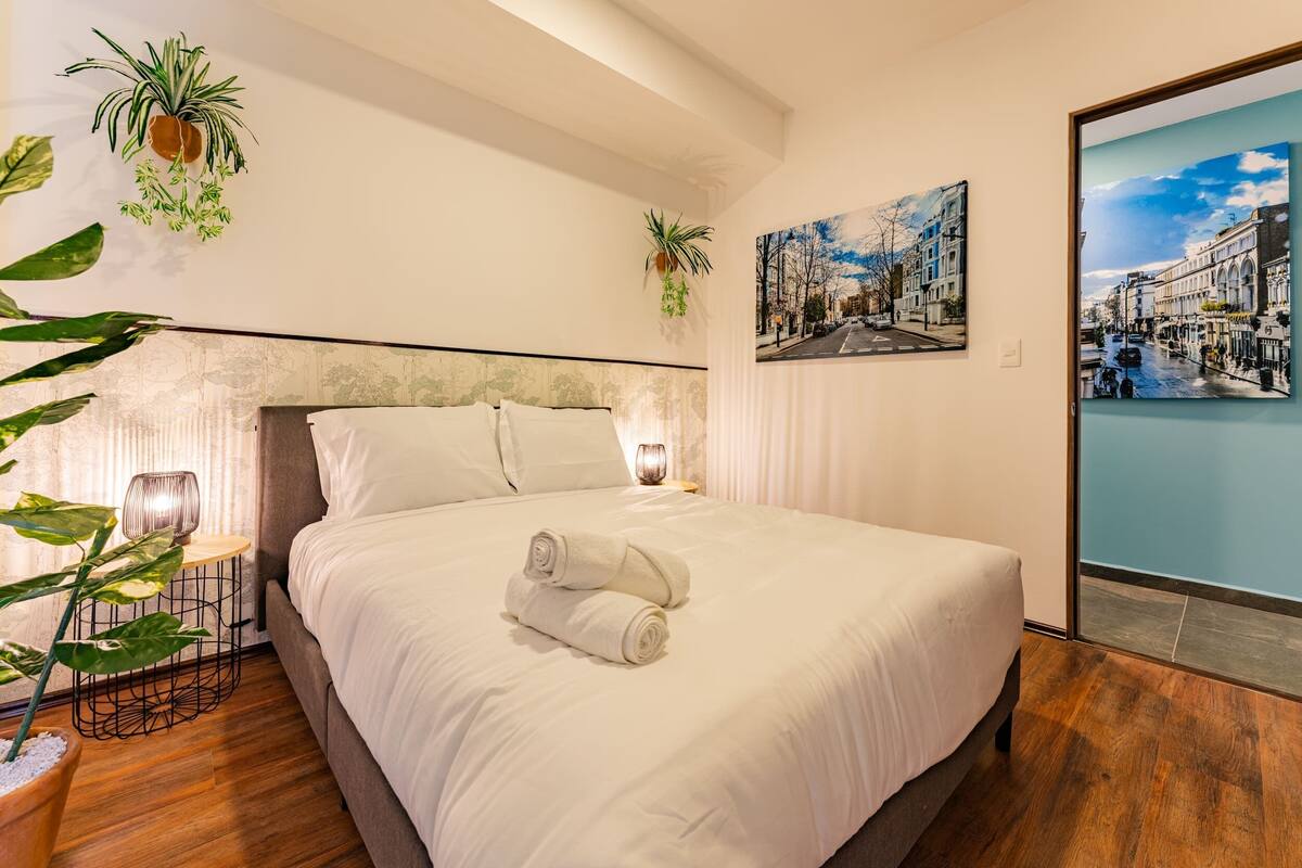 A neatly made bed with white linens and a rolled towel, a small side table with a lamp, and two wall-mounted plants.