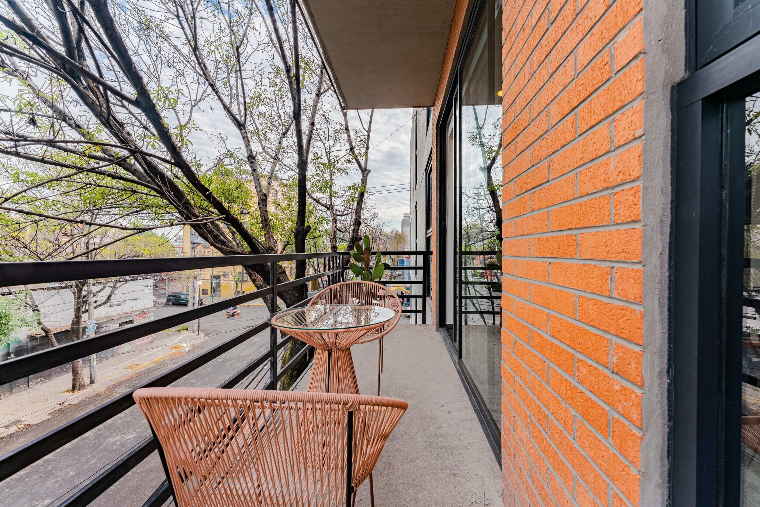 A balcony with wicker chairs and a glass table, overlooking a street with trees and buildings.