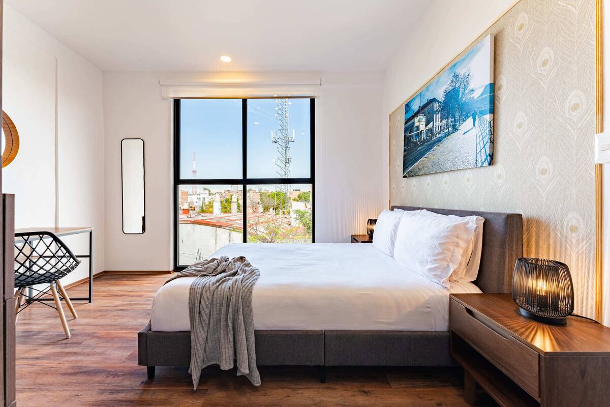 A modern hotel room with a large bed, a desk, and a view of a cityscape through floor-to-ceiling windows.