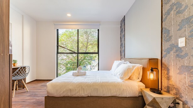 A modern bedroom with a large window, a bed, a desk, and a unique wall lamp. - Doctores Apartments by VH (Mexico City)