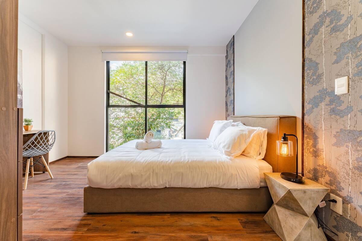 A modern bedroom with a large window, a bed, a desk, and a unique wall lamp.