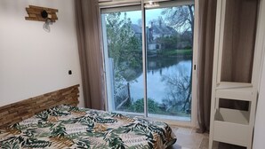 Een slaapkamer met een bed, een houten hoofdeinde, een raam met gordijnen en uitzicht op een meer en bomen.