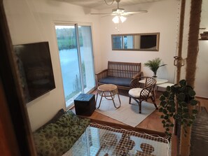 Une pièce confortable avec un canapé, deux fauteuils, une petite table, un téléviseur, un ventilateur de plafond et une grande fenêtre donnant sur l’extérieur.