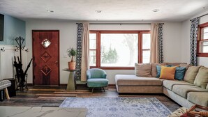 Un salon avec un canapé, un fauteuil et une petite table sur laquelle est posée une plante. On y trouve une porte en bois avec un miroir, ainsi qu'un porte-manteau avec des parapluies.