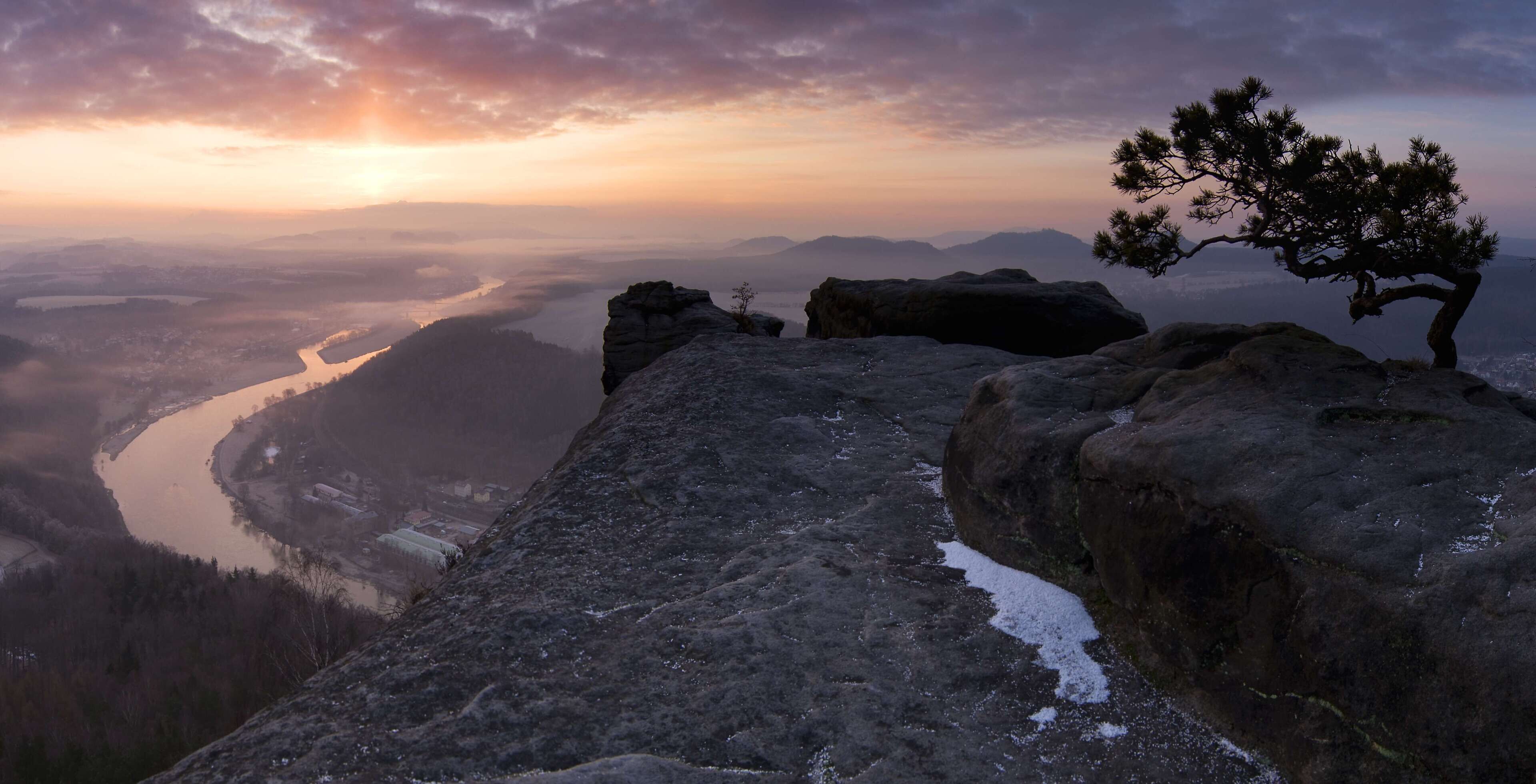 Mirante rochoso com um pinheiro, dando vista para um rio e montanhas ao longe.