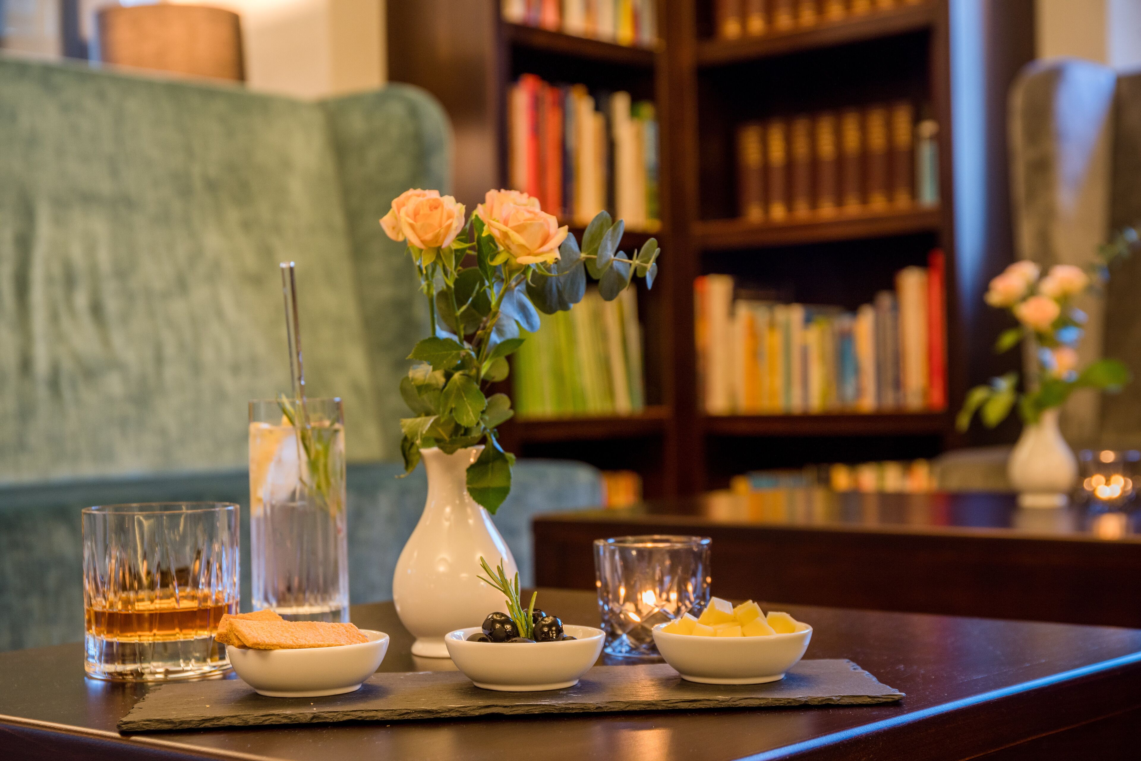 A table with a glass of whiskey, a bowl of olives, a bowl of fruit, and a vase with flowers.