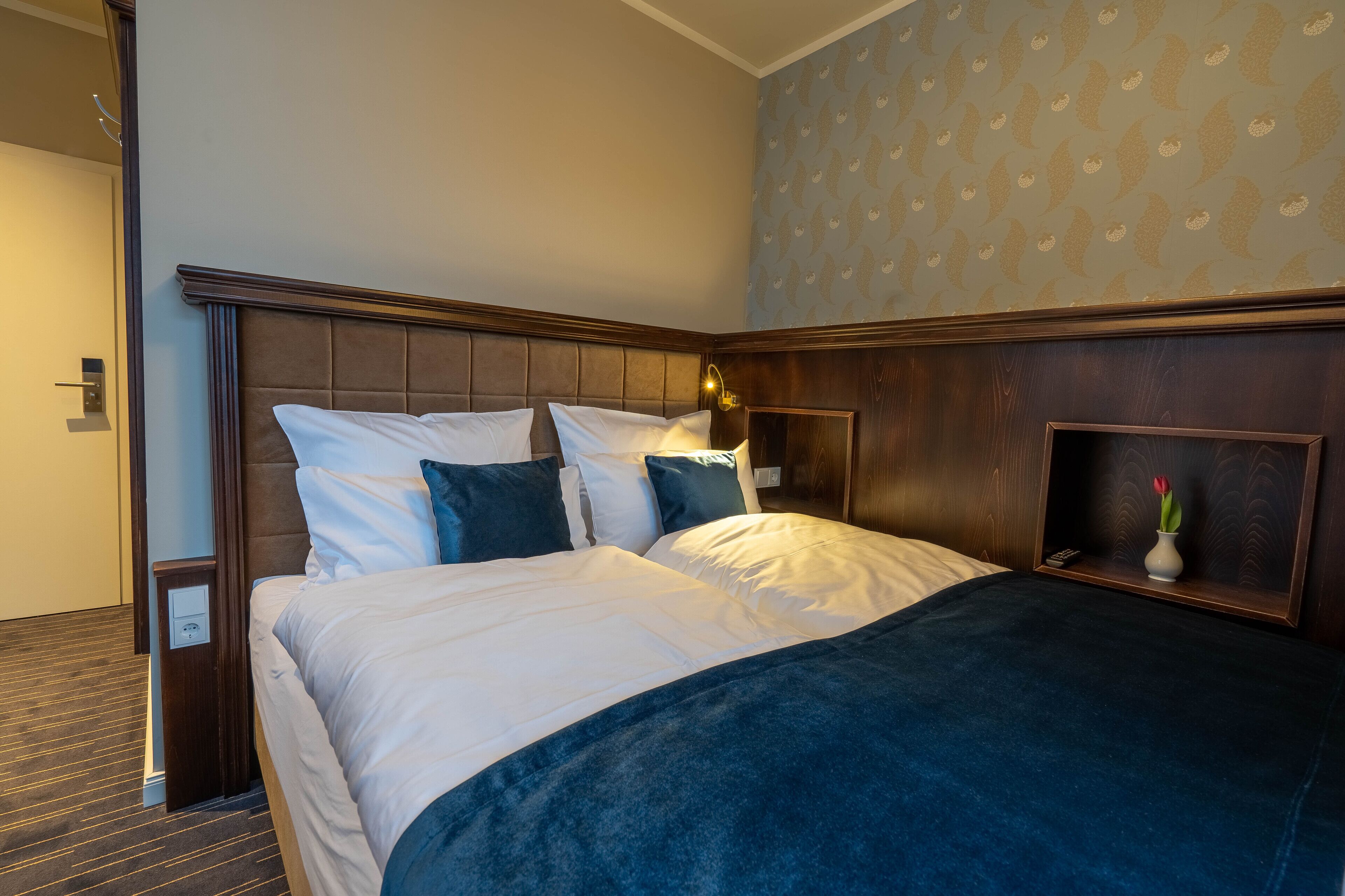 Una cama bien hecha con ropa de cama blanca y azul, una mesita de noche con una lámpara y una pared con papel tapiz estampado.