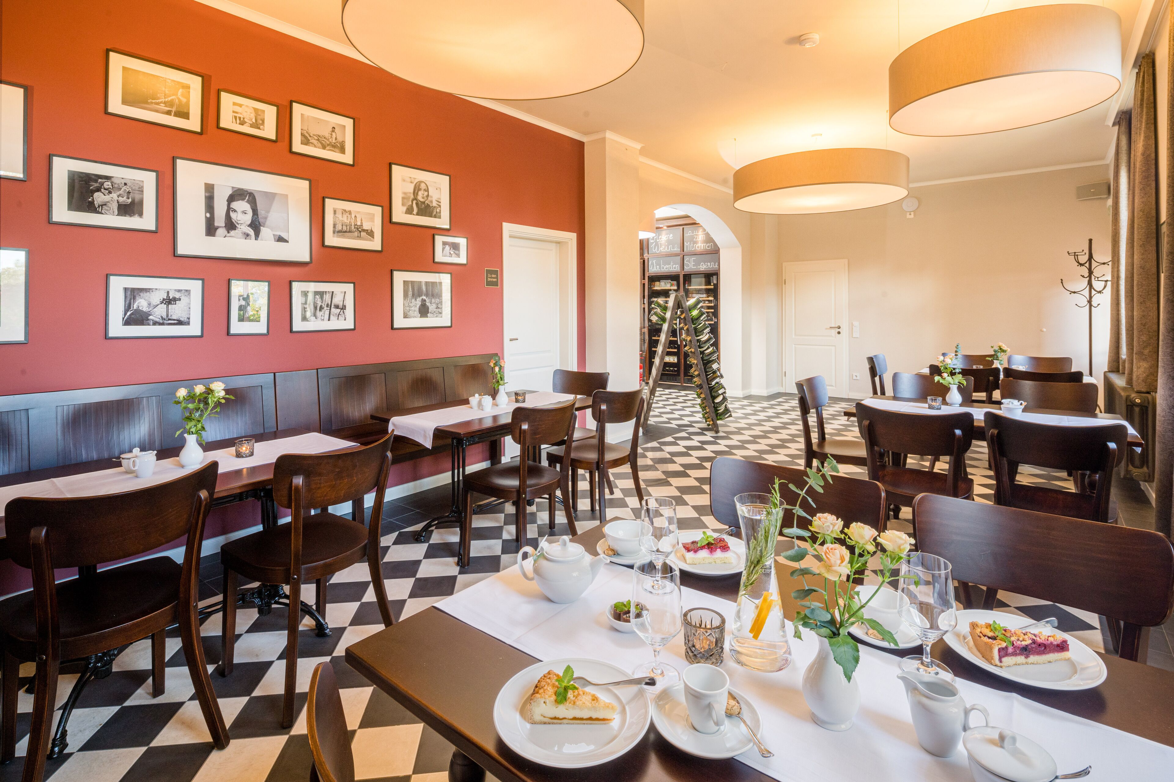 A restaurant with checkered flooring, wooden tables, and chairs. There are framed pictures on the wall, a ladder with bottles, and a white door in the background.