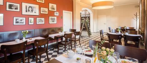 A restaurant with checkered flooring, wooden tables, and chairs. There are framed pictures on the wall, a ladder with bottles, and a white door in the background.