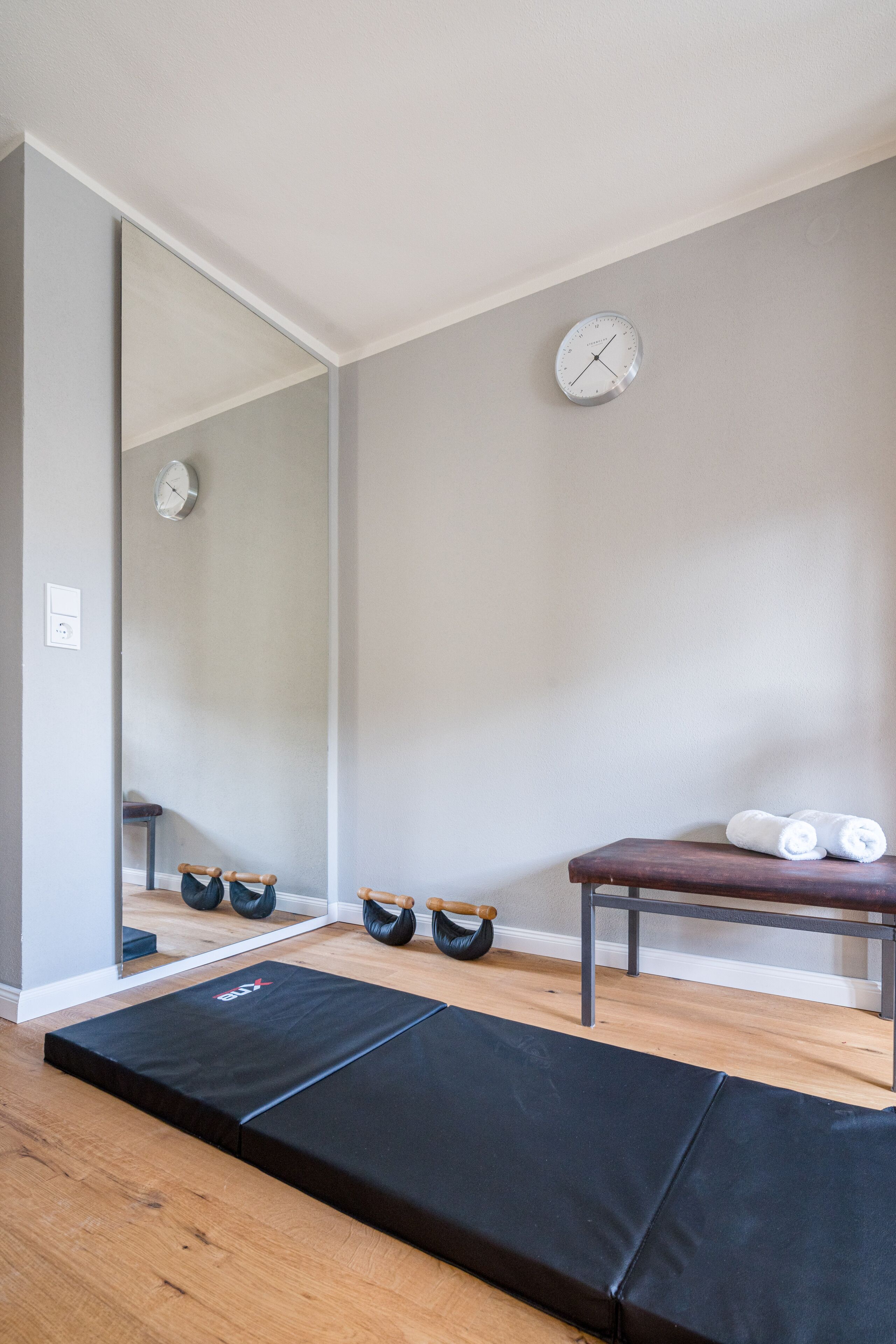 A room with a wooden floor, a black exercise mat, a bench, and a mirror on the wall.