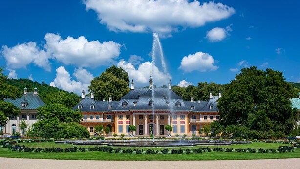 Un edificio grande e histórico con una fuente en primer plano, rodeado de jardines cuidados y árboles.