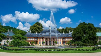 A large, historic building with a fountain in front, surrounded by well-maintained gardens and trees.