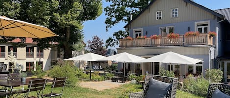 An outdoor seating area with tables and chairs, surrounded by greenery and a large building with a balcony.