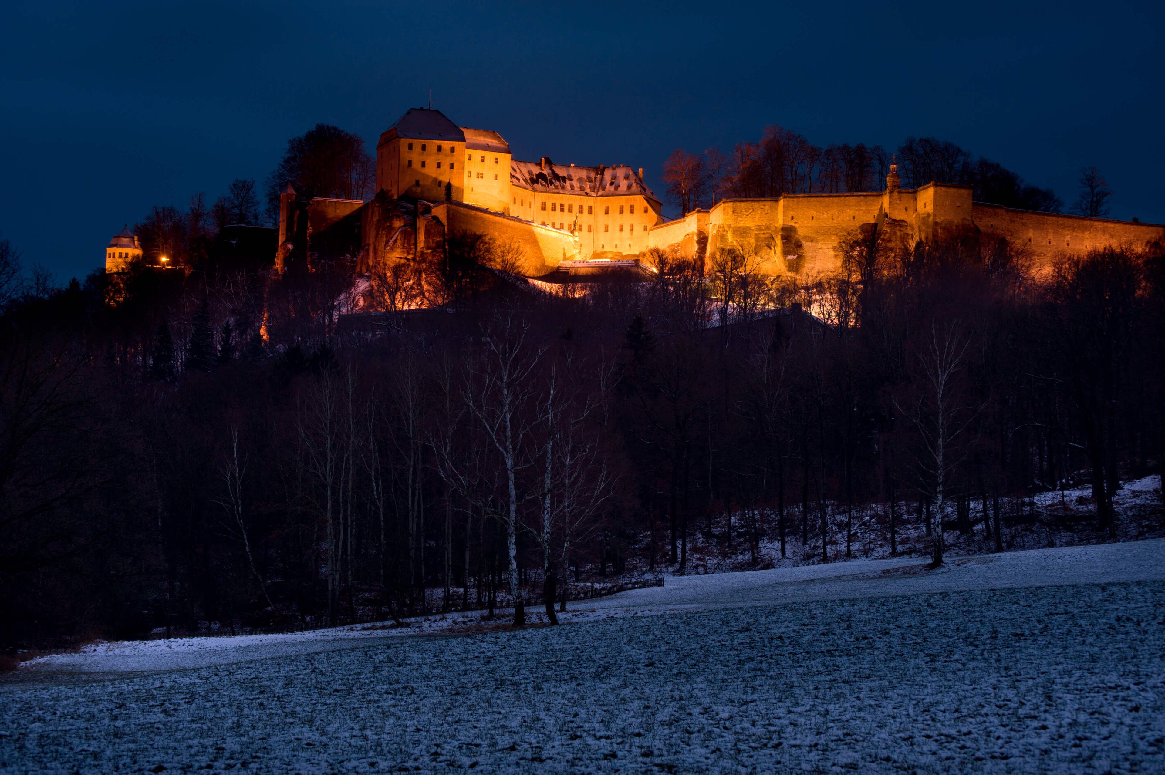 A historic castle with a fortified wall, a large central building, and a smaller structure to the left.