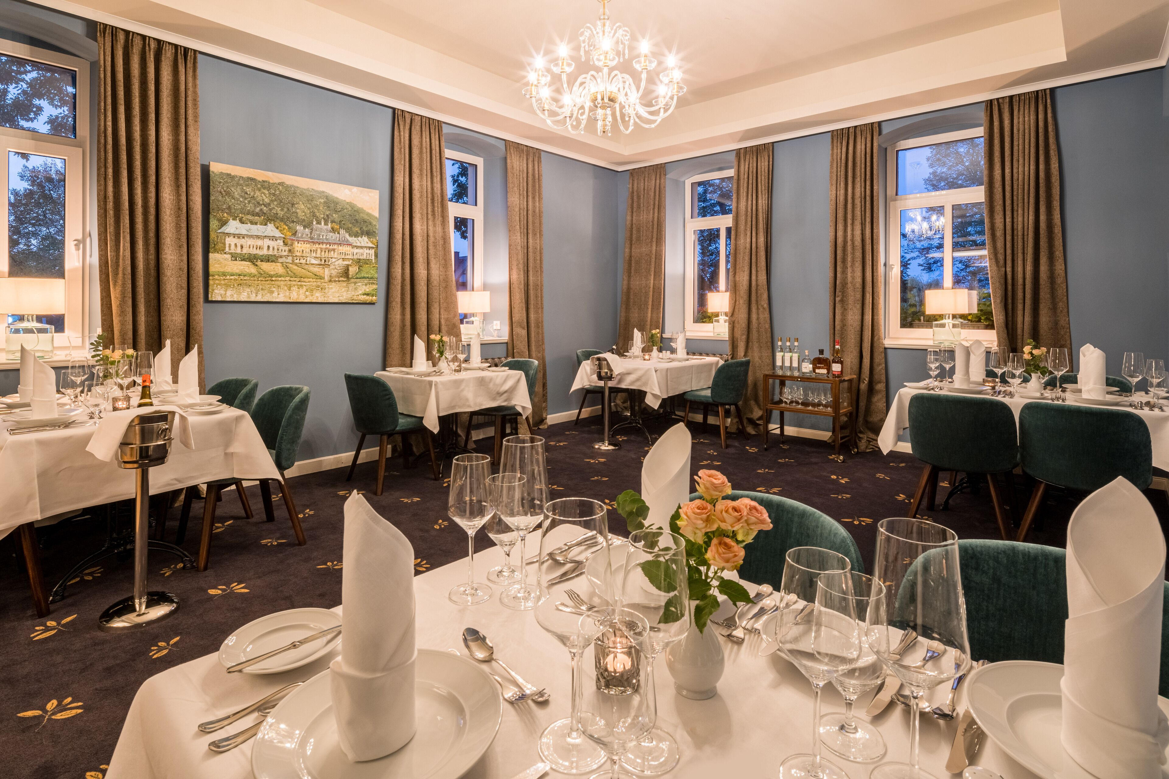 A dining room with multiple tables set for a meal, featuring white tablecloths, green chairs, and a chandelier.