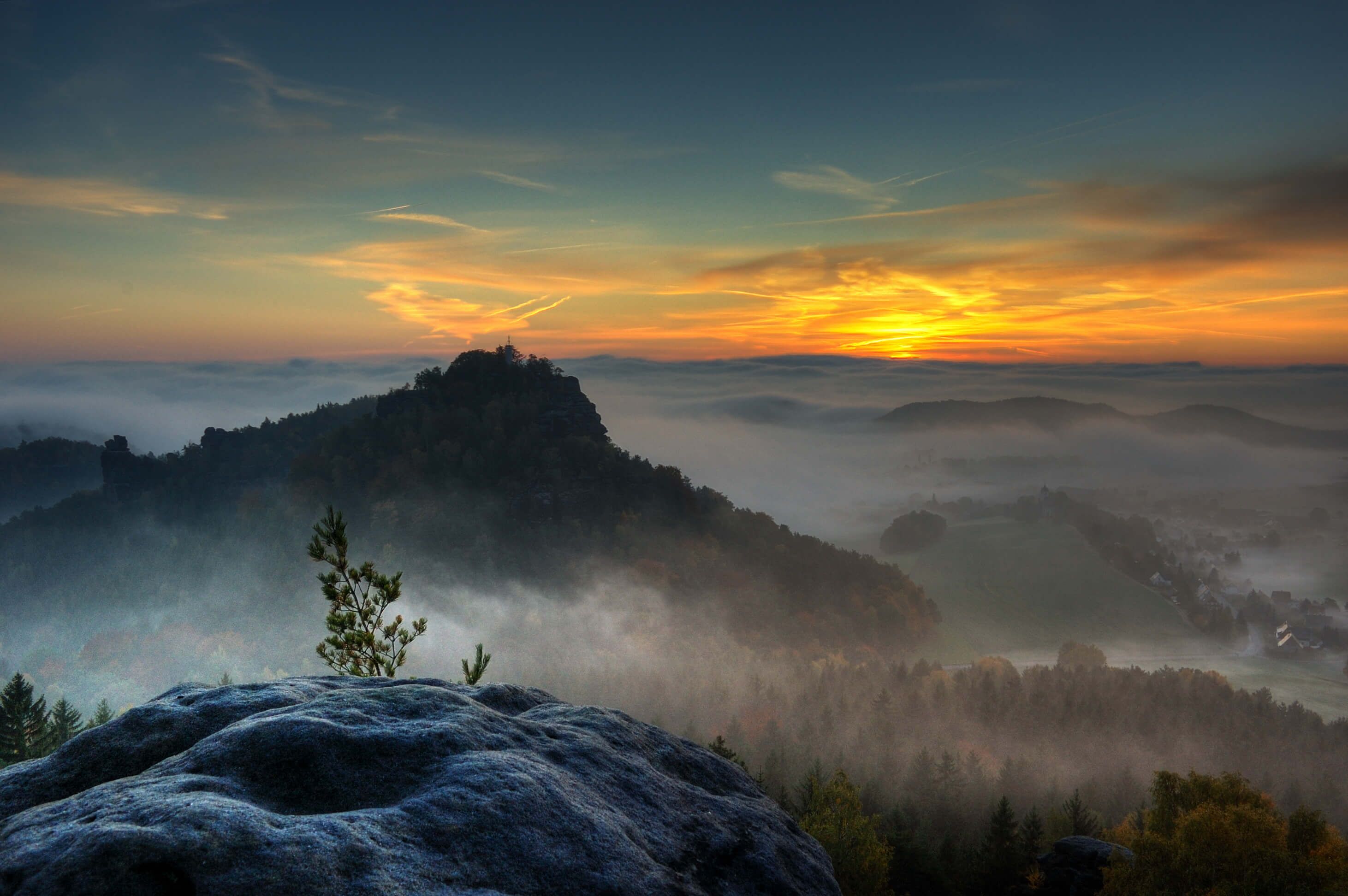 A mountainous landscape with a sunrise or sunset, featuring a prominent rock formation in the foreground and a village in the valley below.