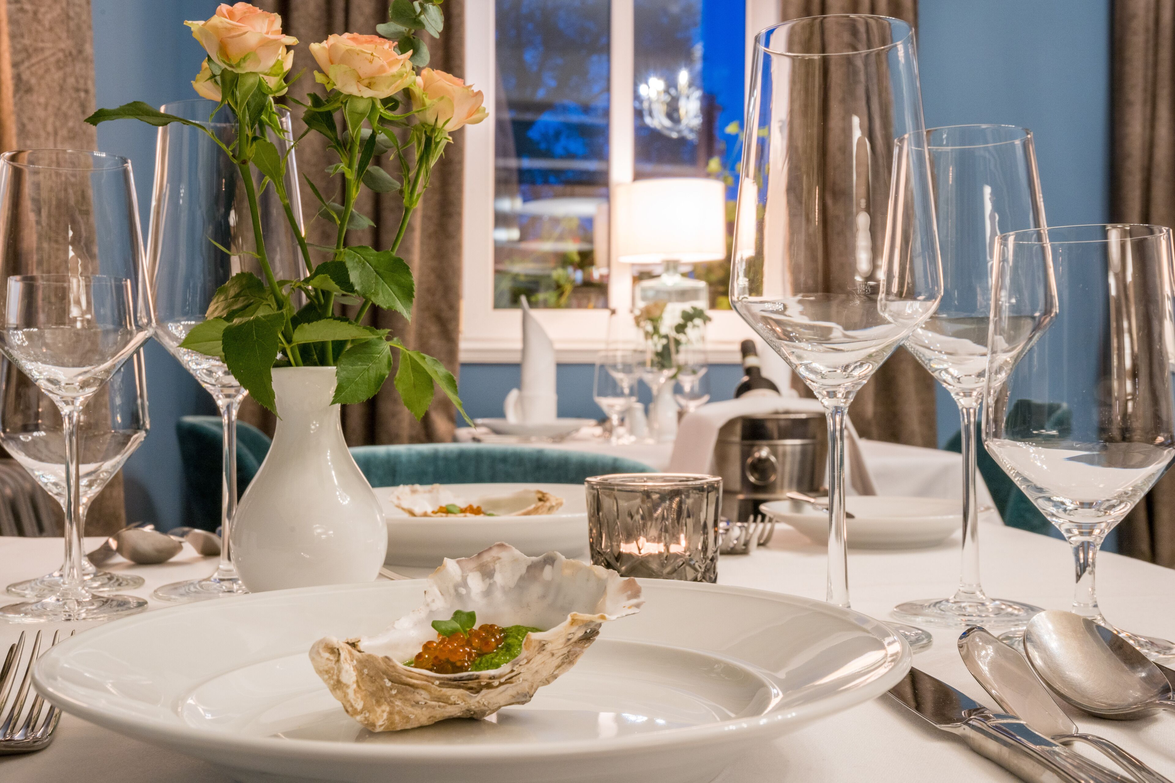 A dining table set with multiple wine glasses, a vase with roses, and a plate with an oyster appetizer.