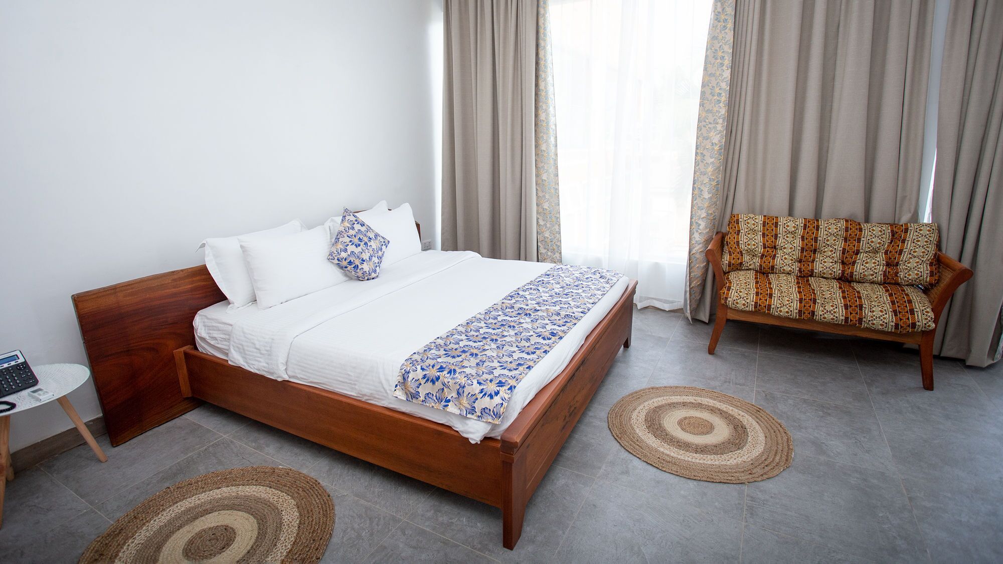 Habitación de hotel ordenada con una cama de madera, un sofá estampado y dos alfombras tejidas en el suelo.