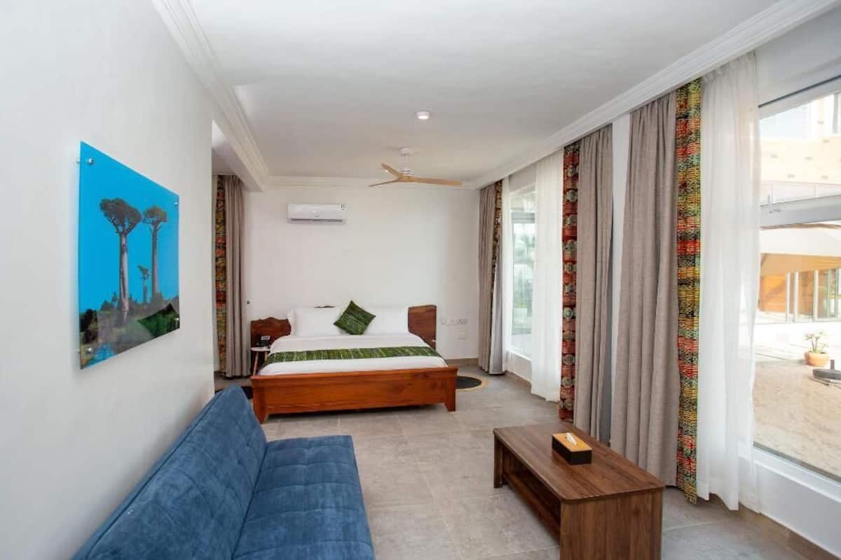 Una habitación de hotel con una cama, un sofá azul, una mesita de madera, un ventilador de techo y un ventanal con cortinas.