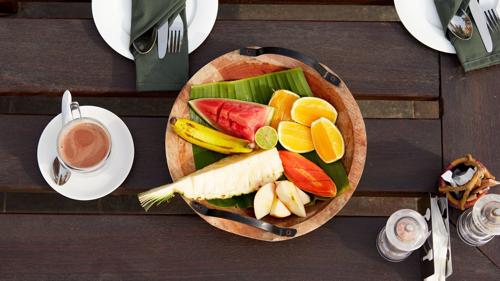 A wooden table with a fruit and vegetable platter, a cup of coffee, and a napkin.