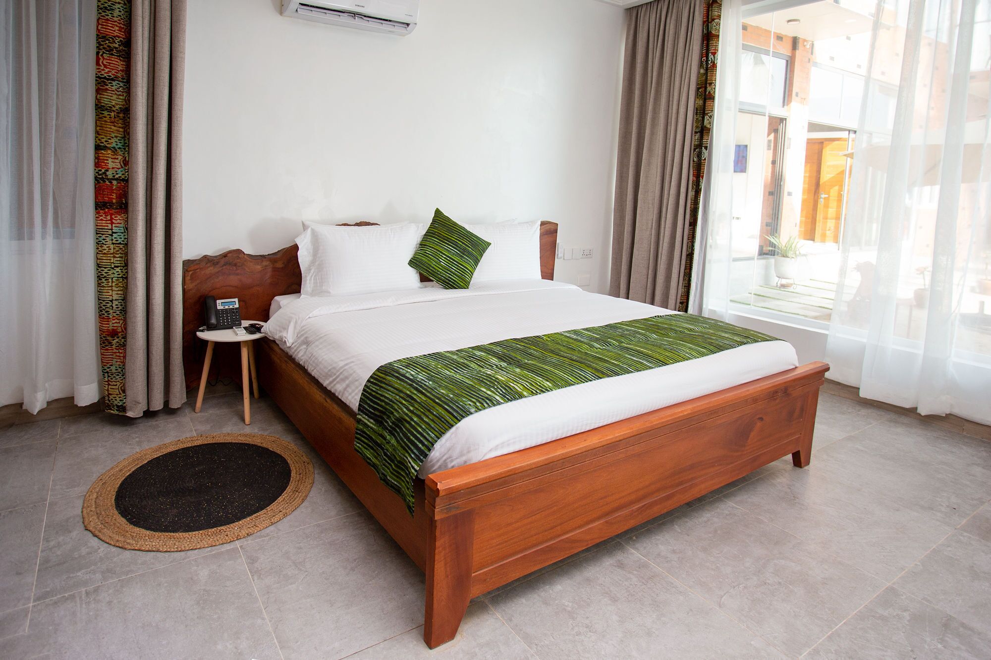 A hotel room with a wooden bed, white bedding, a small bedside table with a phone, and a window with curtains.