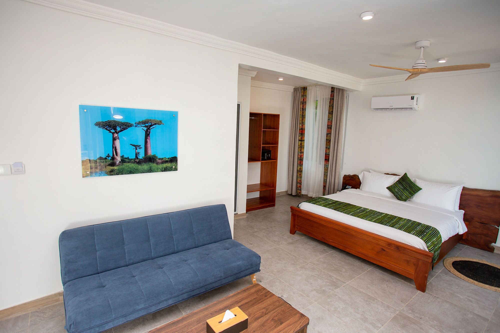 A bedroom with a bed, a blue sofa, a wooden coffee table, a wardrobe, and an air conditioning unit.