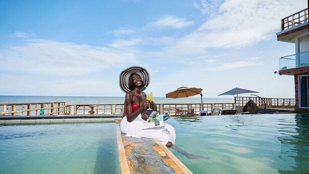 A person sitting on a wooden board in a pool with a view of the ocean and a beach. - La Villa Aphro Ghana (Kokrobite)