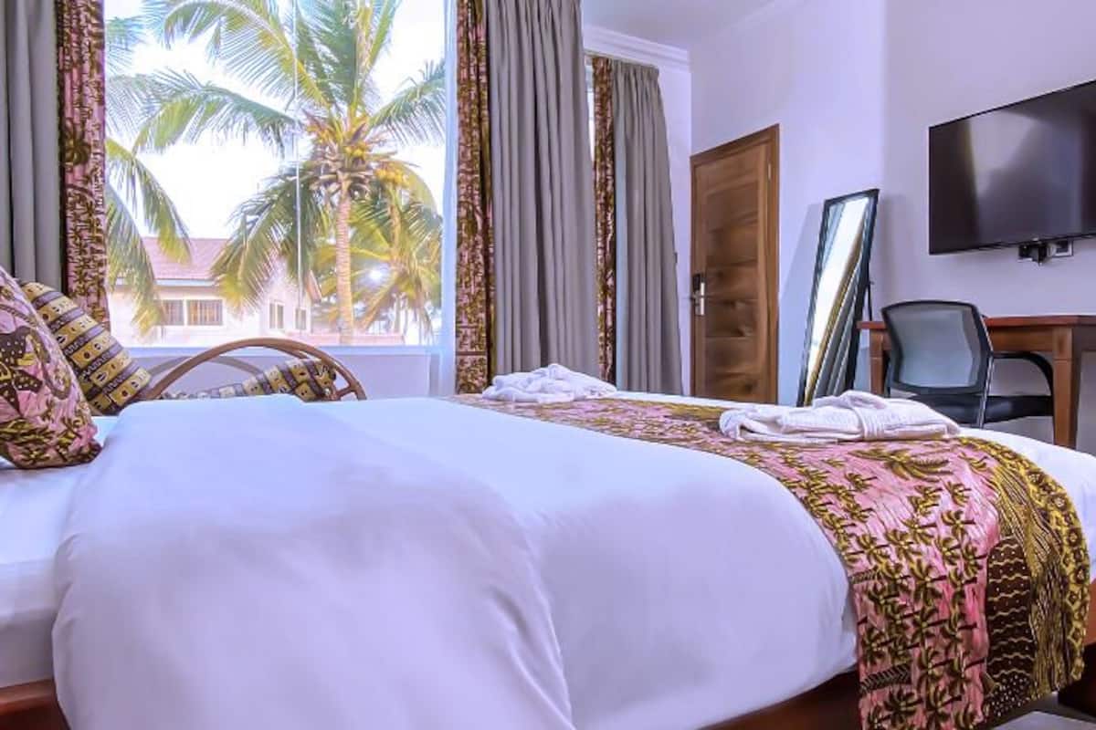 Habitación de hotel con cama, televisor, escritorio y vista a palmeras.