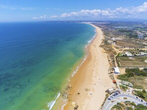 Luftaufnahme einer Küstenlandschaft mit Sandstrand, einer Straße mit Fahrzeugen und Gebäuden in der Ferne.