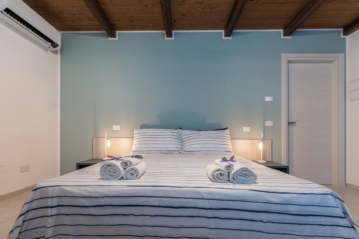 A hotel room with a bed, two bedside tables, a nightstand, an air conditioning unit, and a door.