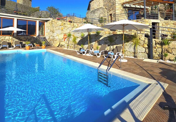 A swimming pool with lounge chairs and umbrellas, stone walls, and a clear blue sky.