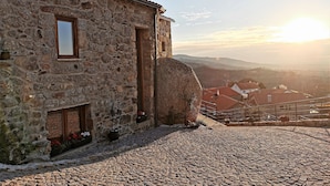 A stone building with a rustic appearance, a cobblestone ground, and a view of a valley with houses in the background. - CASA DO PENEDO CASTLE HOUSE (Linhares da Beira)