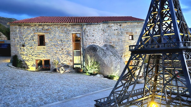 A stone building with a red-tiled roof and a metal structure resembling the Eiffel Tower in the foreground. - CASA DO PENEDO CASTLE HOUSE (Linhares da Beira)