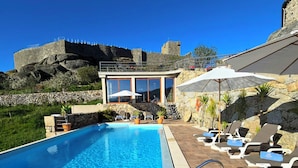 A swimming pool with clear blue water, surrounded by lounge chairs and umbrellas, with a stone building and a castle in the background. - CASA DO PENEDO CASTLE HOUSE (Linhares da Beira)