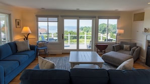En rummelig stue med en blå sofa, en grå sofa og et hvidt sofabord. To vinduer med udsigt til havet og grønne omgivelser.