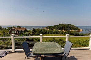 A deck with a table and chairs overlooking a coastal landscape, including a house and a view of the sea. - Panoramic Ocean View; walk to private beach (Gloucester)