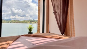 Une chambre avec une grande fenêtre, un lit recouvert d’une couverture beige, une petite plante en pot et une vue sur un lac et des collines.