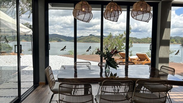 Un coin repas avec une grande table et des chaises en osier, donnant sur un lac et offrant une vue sur des montagnes au loin.