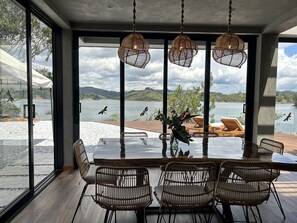 Een eetgedeelte met een grote tafel en rieten stoelen, met uitzicht op een meer met bergen in de verte.