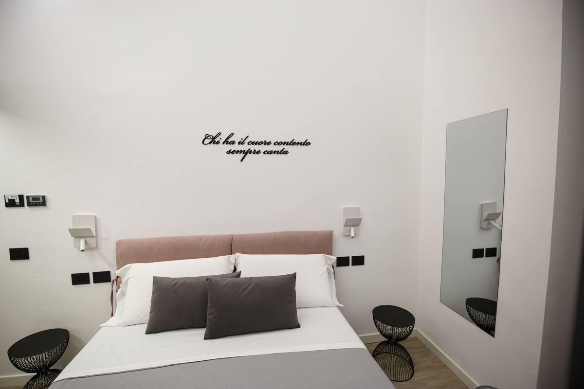 A modern bedroom with a bed, bedside tables, a wall-mounted mirror, and a decorative wall quote.